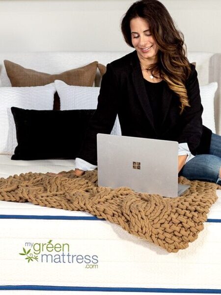 A woman sits on a bed with a laptop on her lap. The mattress has a "My Green Mattress" logo. The bedroom features two wooden nightstands, a throw blanket, and various decorative elements.