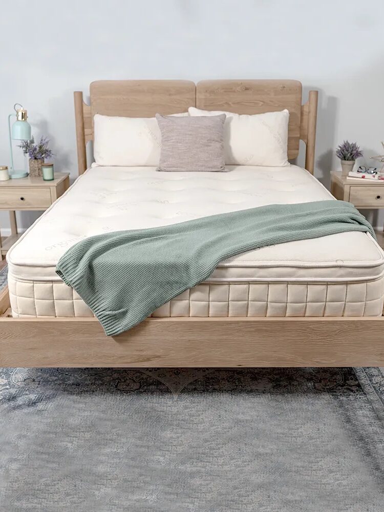 A neatly made bed with a light wood frame and off-white mattress, adorned with a green throw blanket and a beige pillow. Two wooden nightstands with decor are on either side. The room has a light, airy feel.