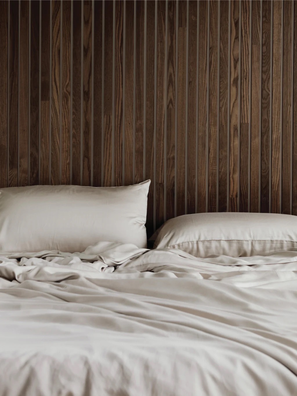 A bed with white sheets and two pillows is in front of a wooden slat wall.