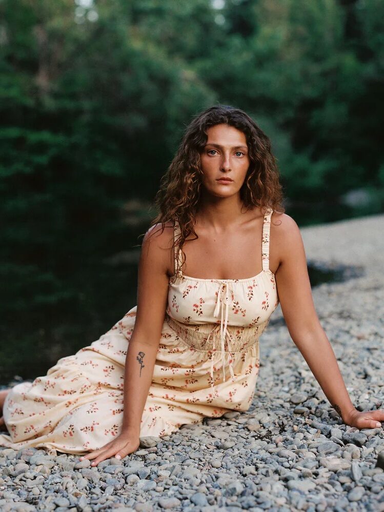 A person with long curly hair wearing a floral dress sits on a pebble-covered surface near a wooded area beside water.