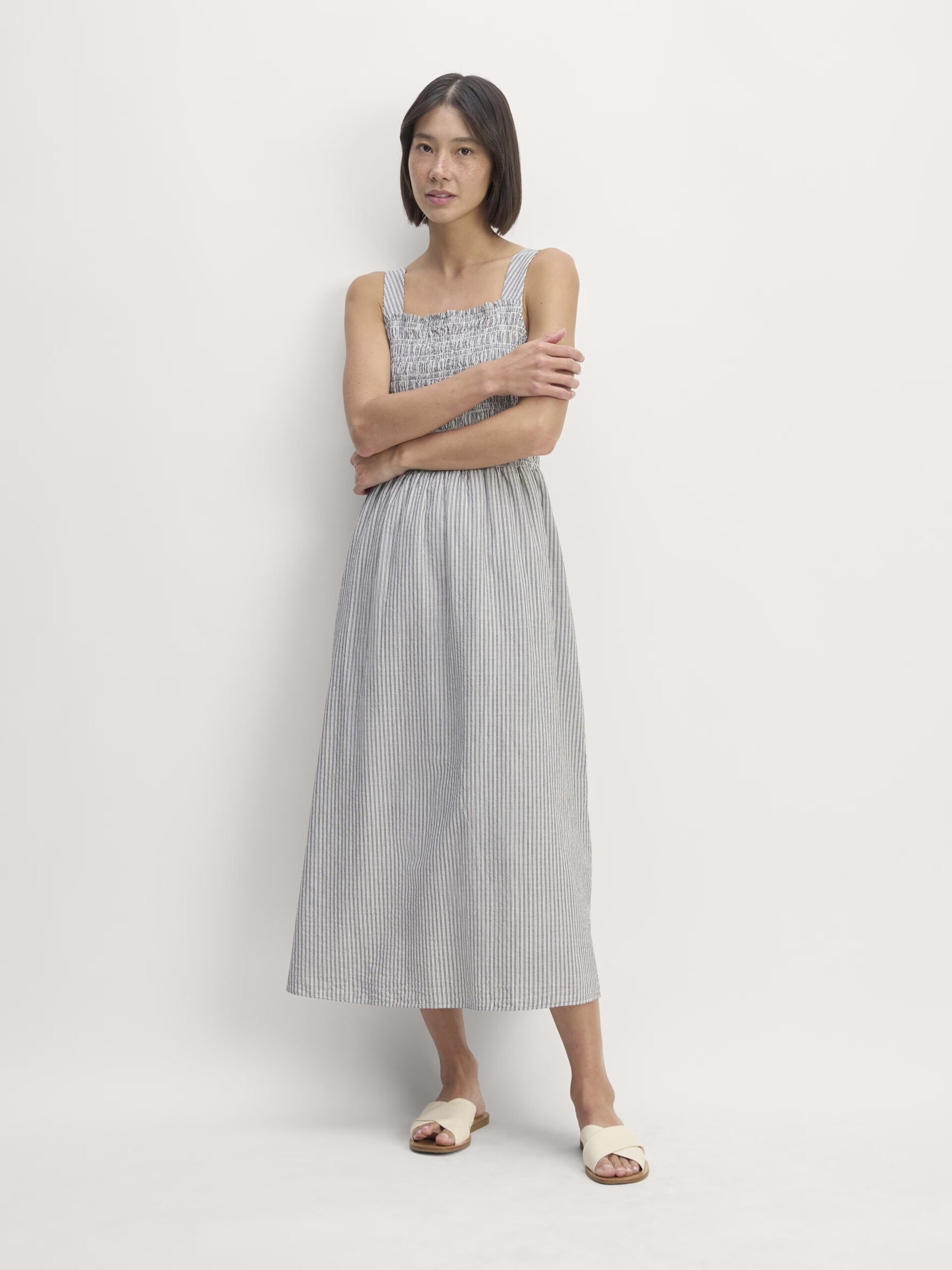 A woman stands against a plain white background, wearing a gray and white striped sleeveless, ankle-length dress and white slide sandals. She has her arms crossed and is looking ahead.