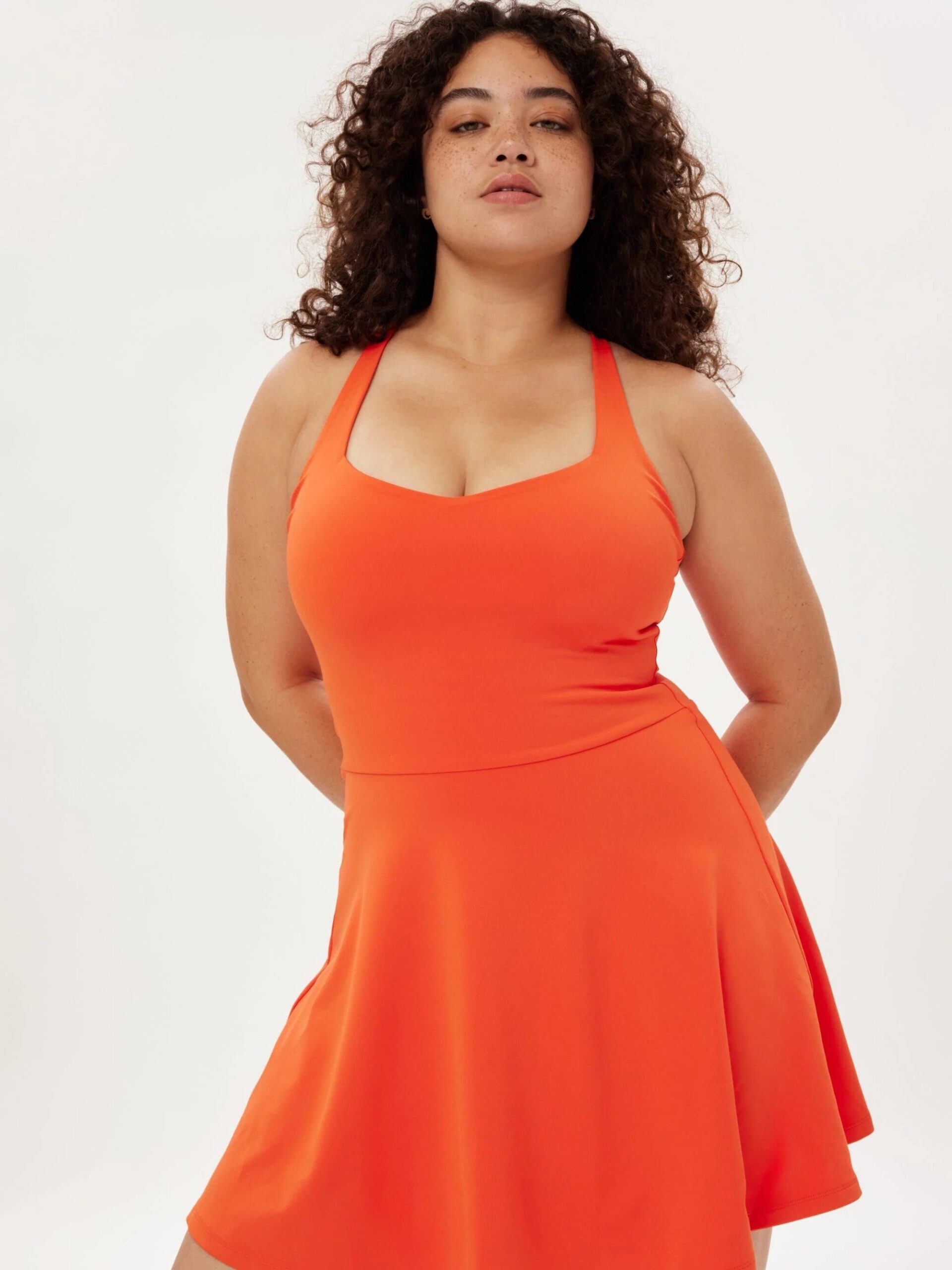 A person with curly hair wearing an orange sleeveless dress stands against a plain background, looking at the camera.