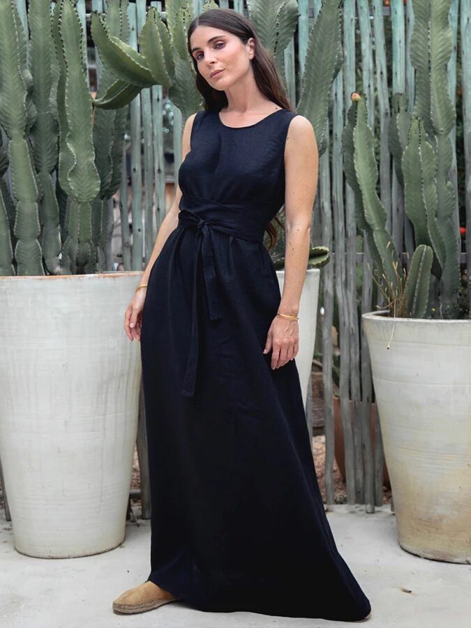 A woman stands in front of tall potted cacti, wearing a sleeveless black dress with a cinched waist and beige slip-on shoes.