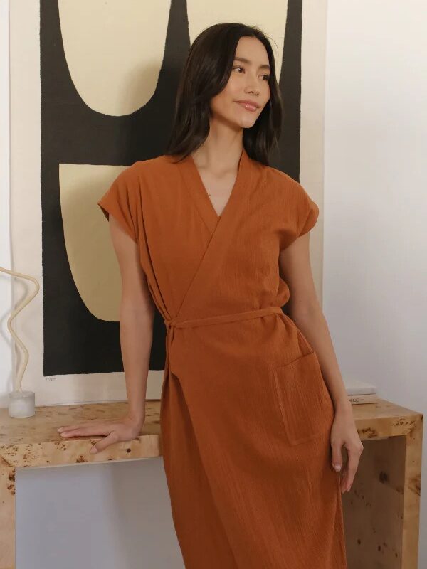 A woman in an orange wrap dress leans against a wooden surface in front of a black and white abstract painting.