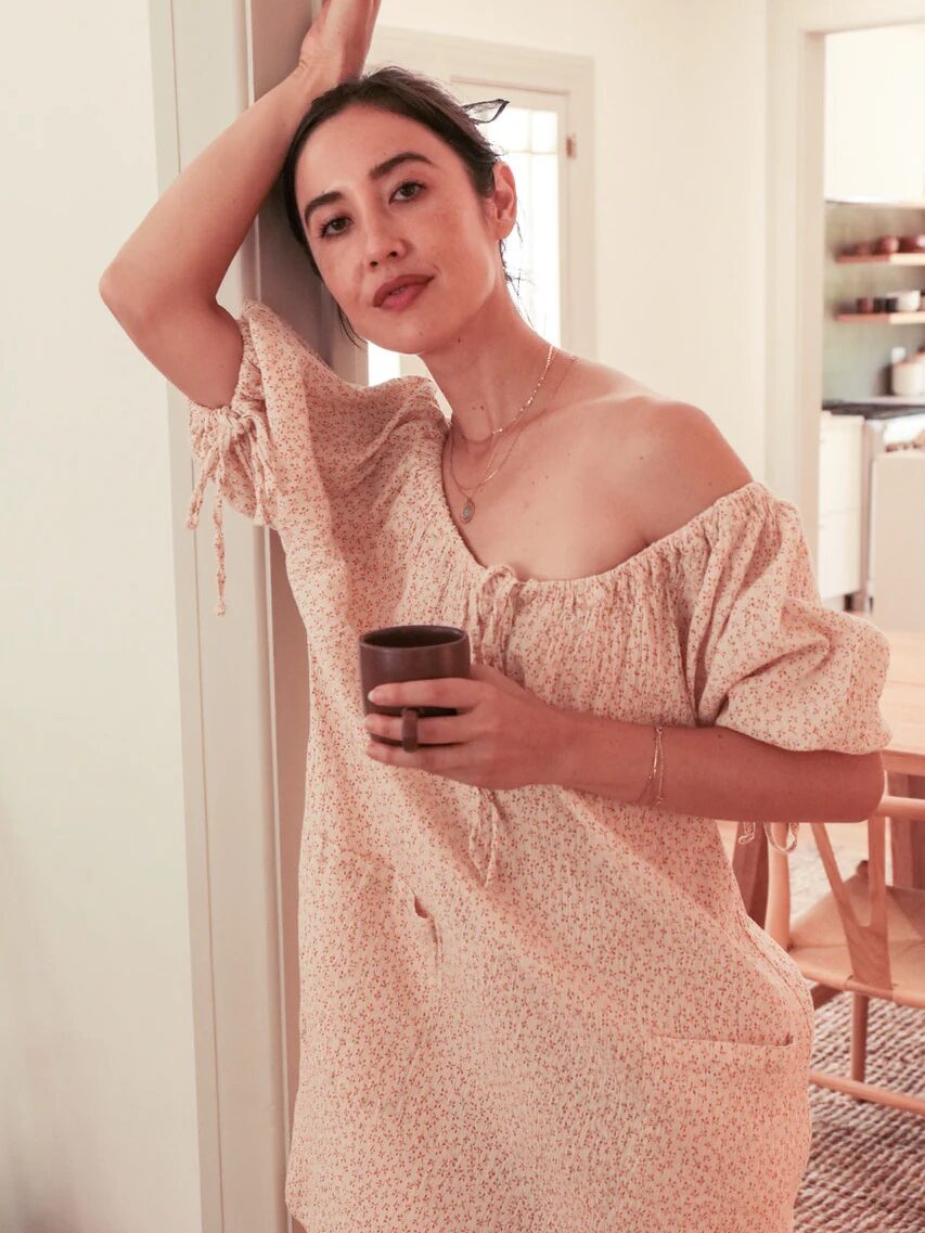 A person with their arm resting on a door frame, wearing an off-the-shoulder dress and holding a cup, stands in a well-lit room. A dining table and chairs are visible in the background.