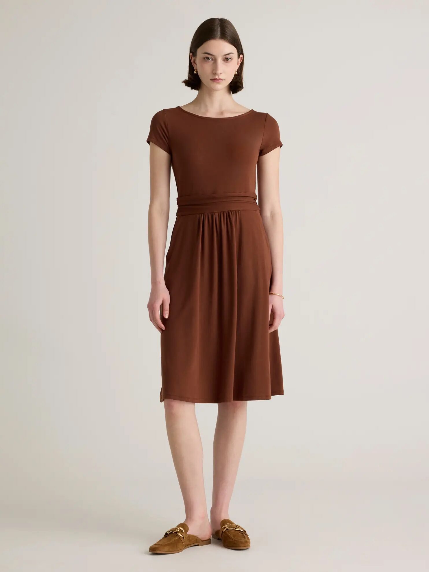 A person stands against a plain background, wearing a short-sleeved brown dress and brown loafers. They have short, straight hair and a neutral expression.