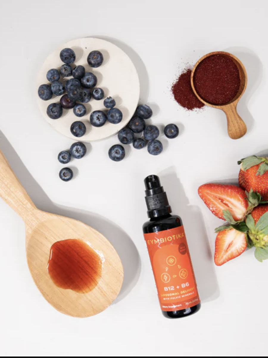 A wooden spoon with liquid, a bottle of Symbiotika B12 + B6, fresh blueberries, a wooden scoop with powdered supplement, and three fresh strawberries on a white surface.