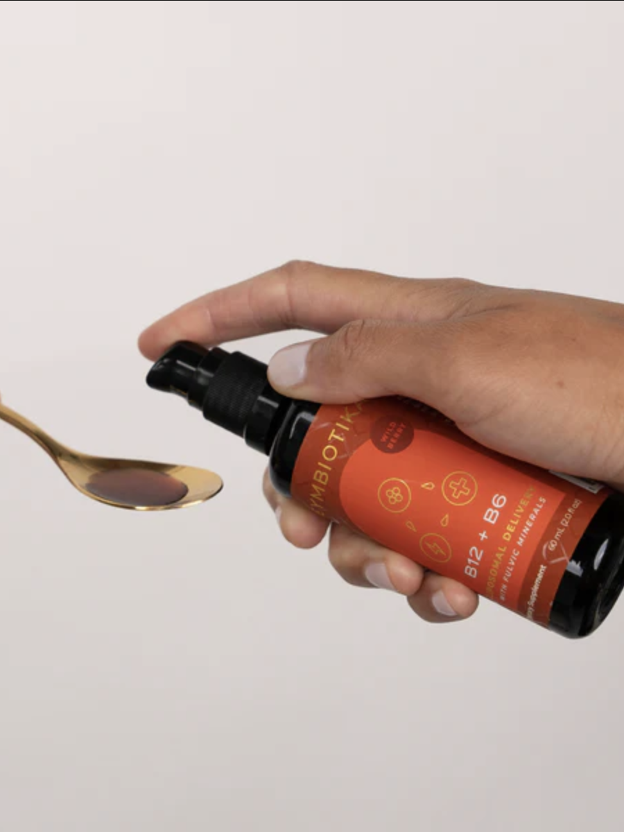 Two hands are pictured; one hand holds a spoon while the other dispenses a liquid supplement from a black bottle with a red label into the spoon.