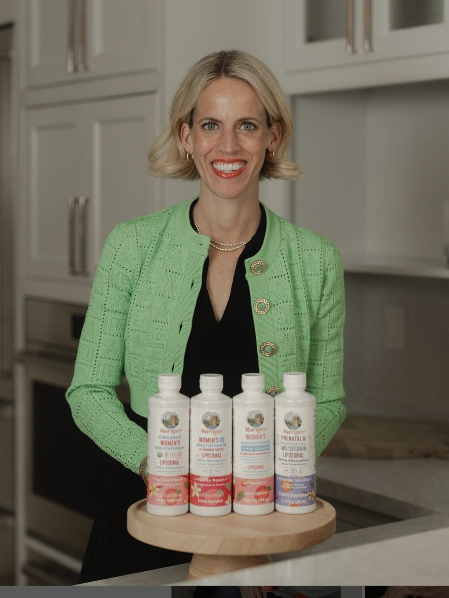 A woman in a green jacket stands in a kitchen behind a round wooden tray displaying four bottles of supplements.