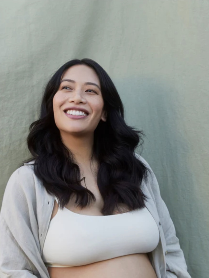 A smiling woman with long dark hair, wearing a light-colored shirt and white top, stands in front of a plain light-colored fabric background.