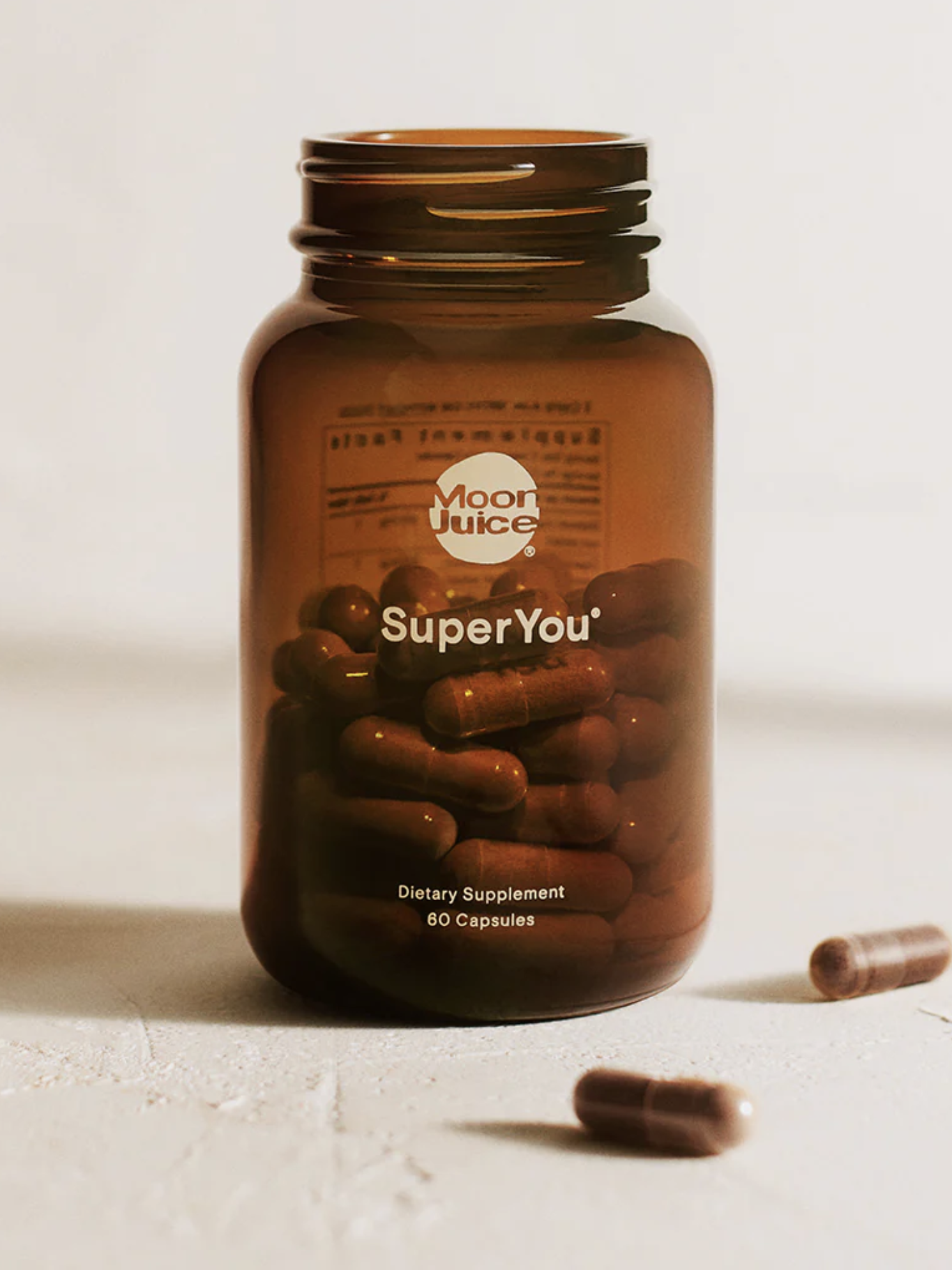 A brown bottle labeled "SuperYou" from Moon Juice, containing dietary supplement capsules, with two capsules outside the bottle.