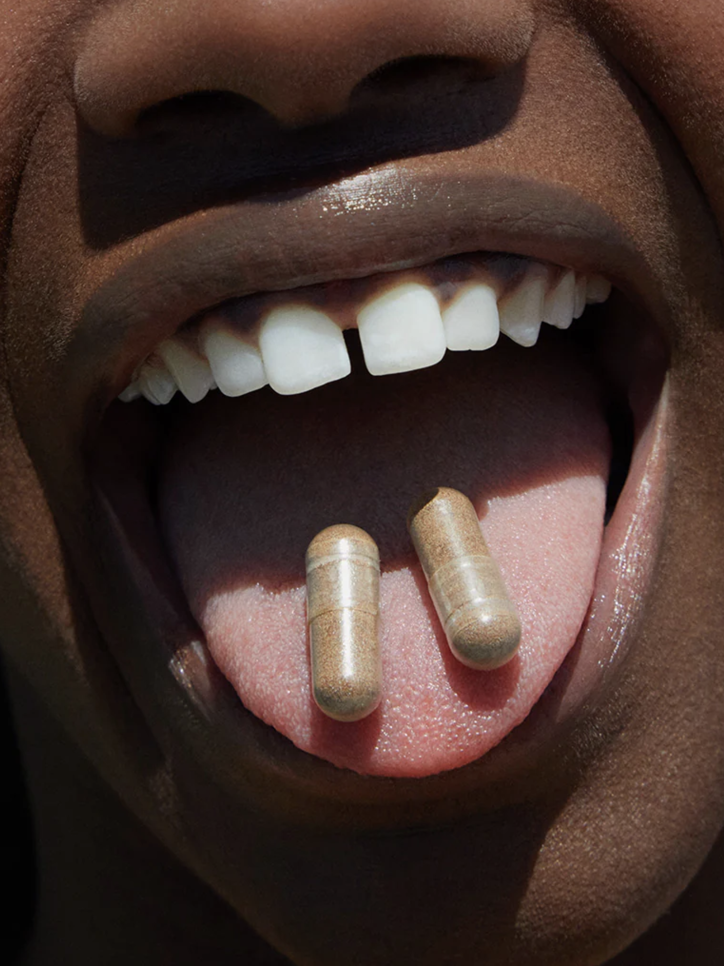 Close-up of a person holding two capsules on their tongue. Their mouth is open, showing their teeth.