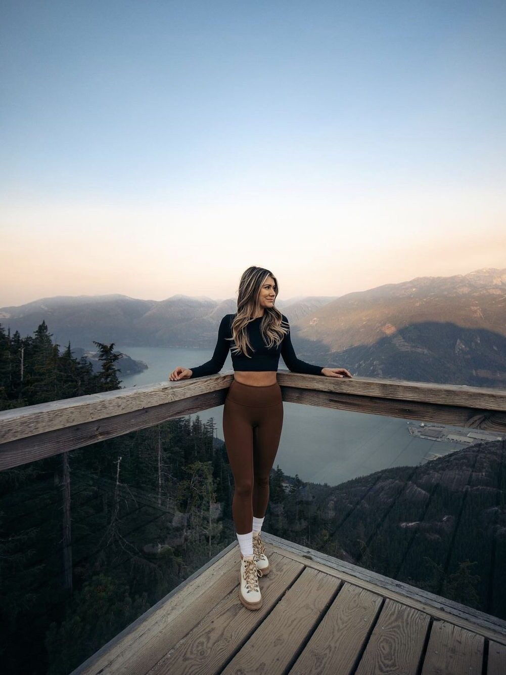 A woman stands on a wooden platform overlooking a vast mountainous landscape with a lake below, under a clear sky at dusk.