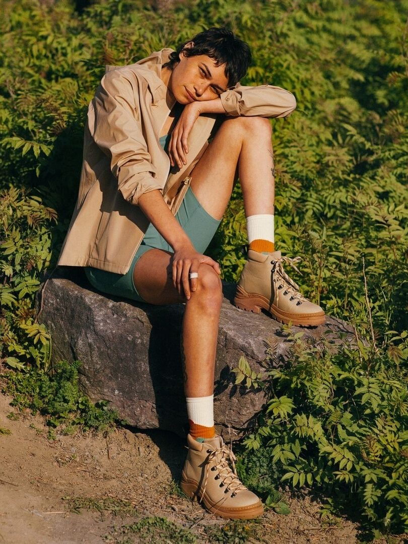 A person sits on a rock, surrounded by greenery, wearing a beige jacket, green shorts, beige boots, and orange socks.