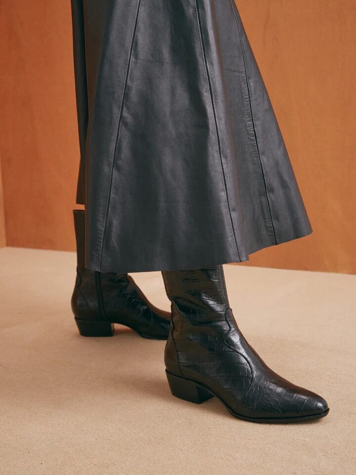 A person wearing black knee-high leather boots with low heels and a dark, long leather coat. The background is a room with a beige floor and wooden wall.