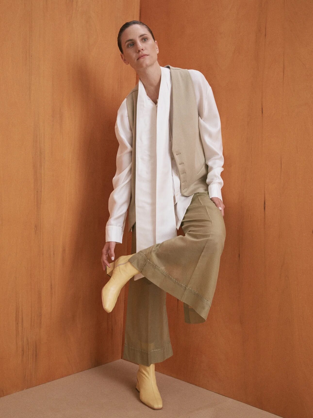 Person in a white shirt, beige vest, and wide-legged khaki pants standing against a wooden wall, with one leg slightly raised and yellow boots visible.