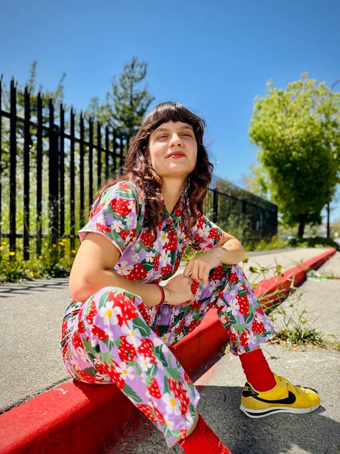 Person sitting on a sidewalk curb wearing a brightly colored floral outfit, red socks, and yellow sneakers. A black metal fence and green trees are visible in the background.