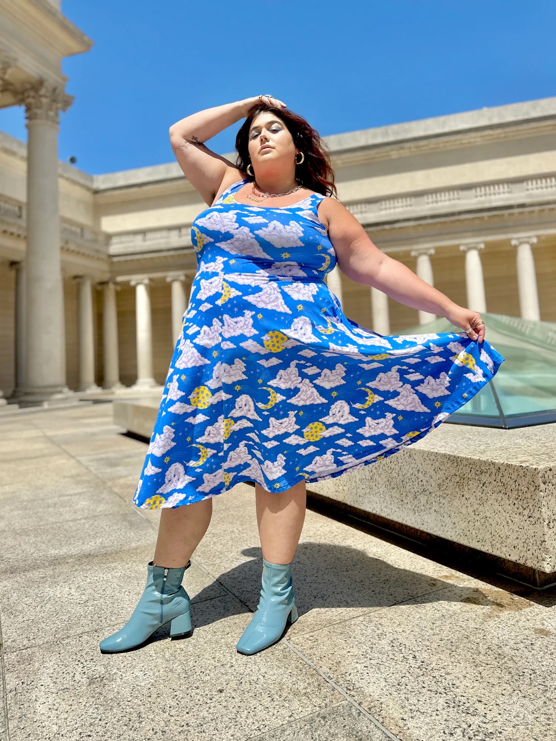 A model wears a bright blue mid length summer dress with a cloud and moon pattern, from Nooworks. 