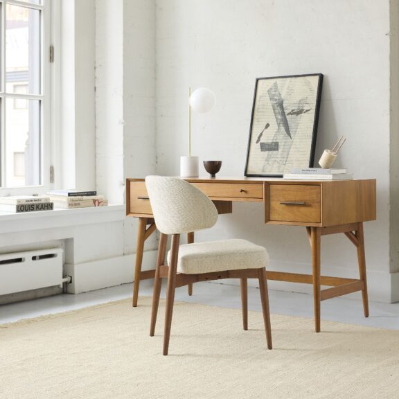 A wooden desk with a modern chair is set in a bright room. On the desk are a small lamp, a cup, and a framed art piece. A stack of books is placed by the window.