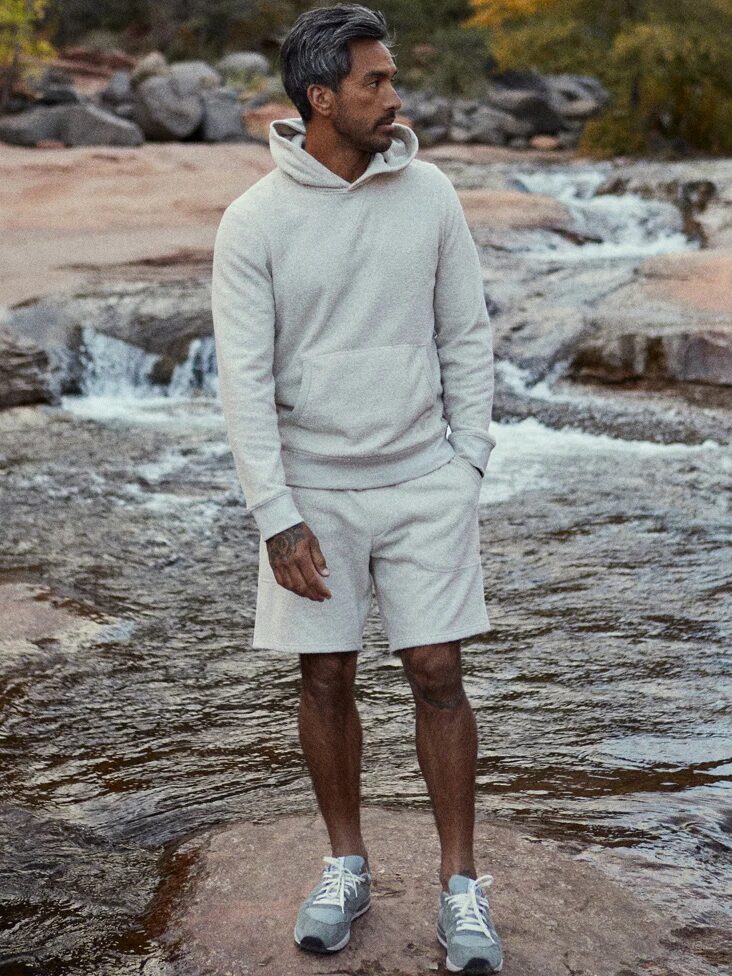 A person in a light-colored hoodie and shorts stands on a rock by a small stream with sparse vegetation in the background.