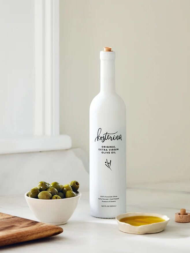 A white bottle of Kosterina original extra virgin olive oil is placed on a kitchen countertop next to a small bowl of green olives, a wooden cutting board, and a small dish with olive oil.