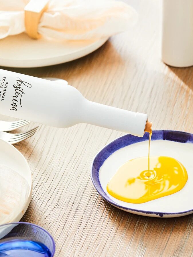 A bottle of orange-colored liquid is being poured onto a small blue-rimmed plate on a wooden surface, surrounded by other plates and a napkin.