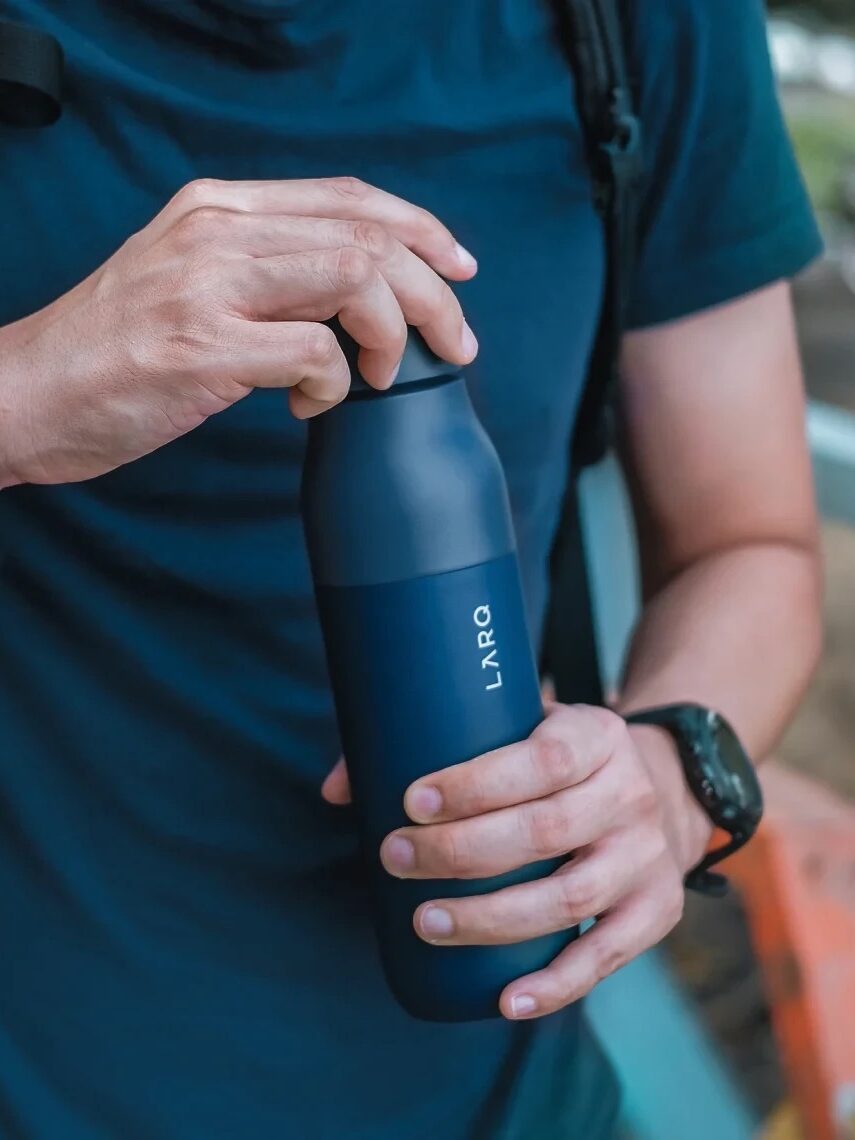 Person in a blue shirt holding and opening a blue LARQ water bottle with both hands. A backpack strap is visible on one shoulder.