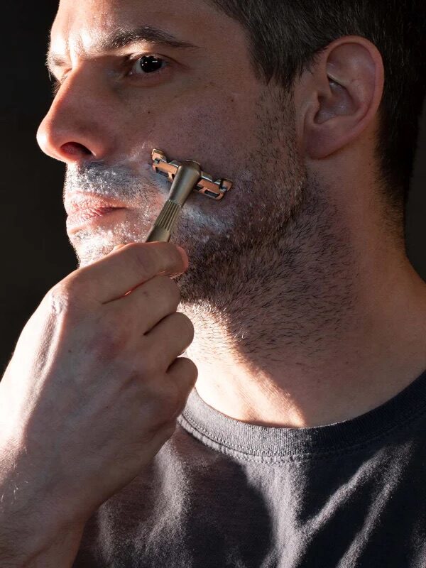 A man with short hair and stubble is shaving his face with a razor. He is wearing a dark-colored t-shirt and has shaving cream applied to his cheek.