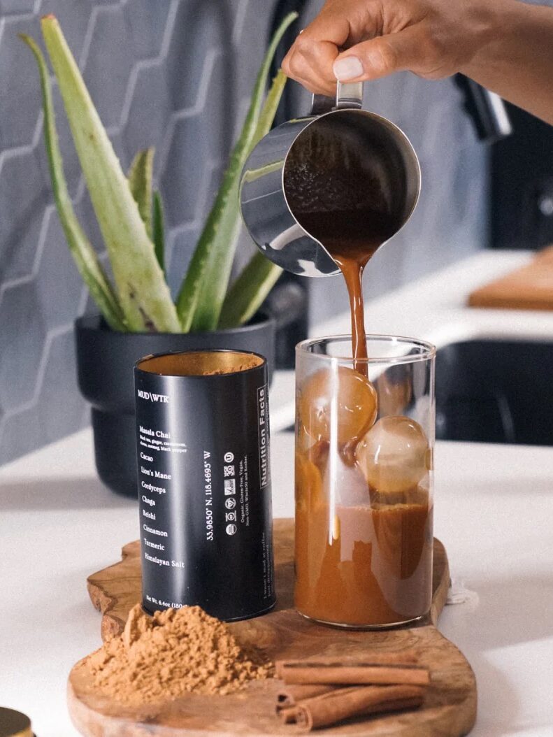 A person pours coffee into a glass with ice spheres. A container of cocoa powder, cinnamon sticks, and a potted aloe vera plant are on a wooden board nearby.