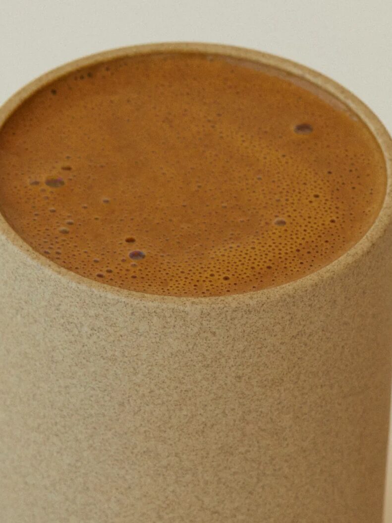 A close-up of a tall, cylindrical cup filled with frothy brown liquid. The cup appears to be made from a plain, textured material.