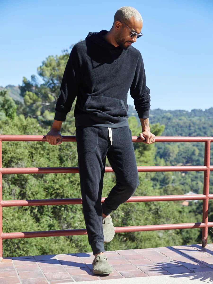 A person in a black hoodie and sweatpants leans on a railing against a backdrop of trees and a clear sky.