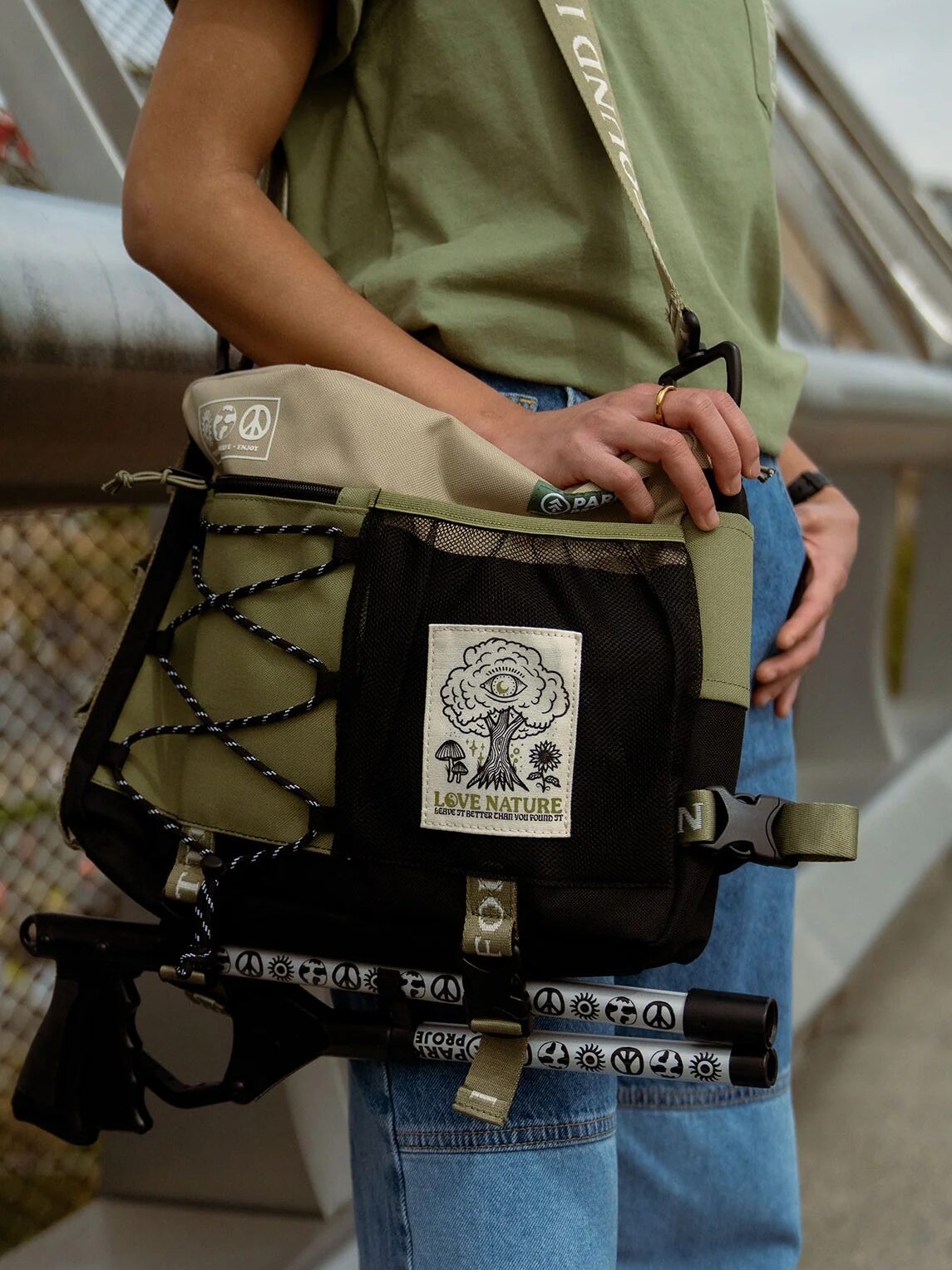 Person wearing a green shirt and blue jeans standing on a bridge, carrying a backpack with a nature-themed patch and two cylindrical items attached to it.