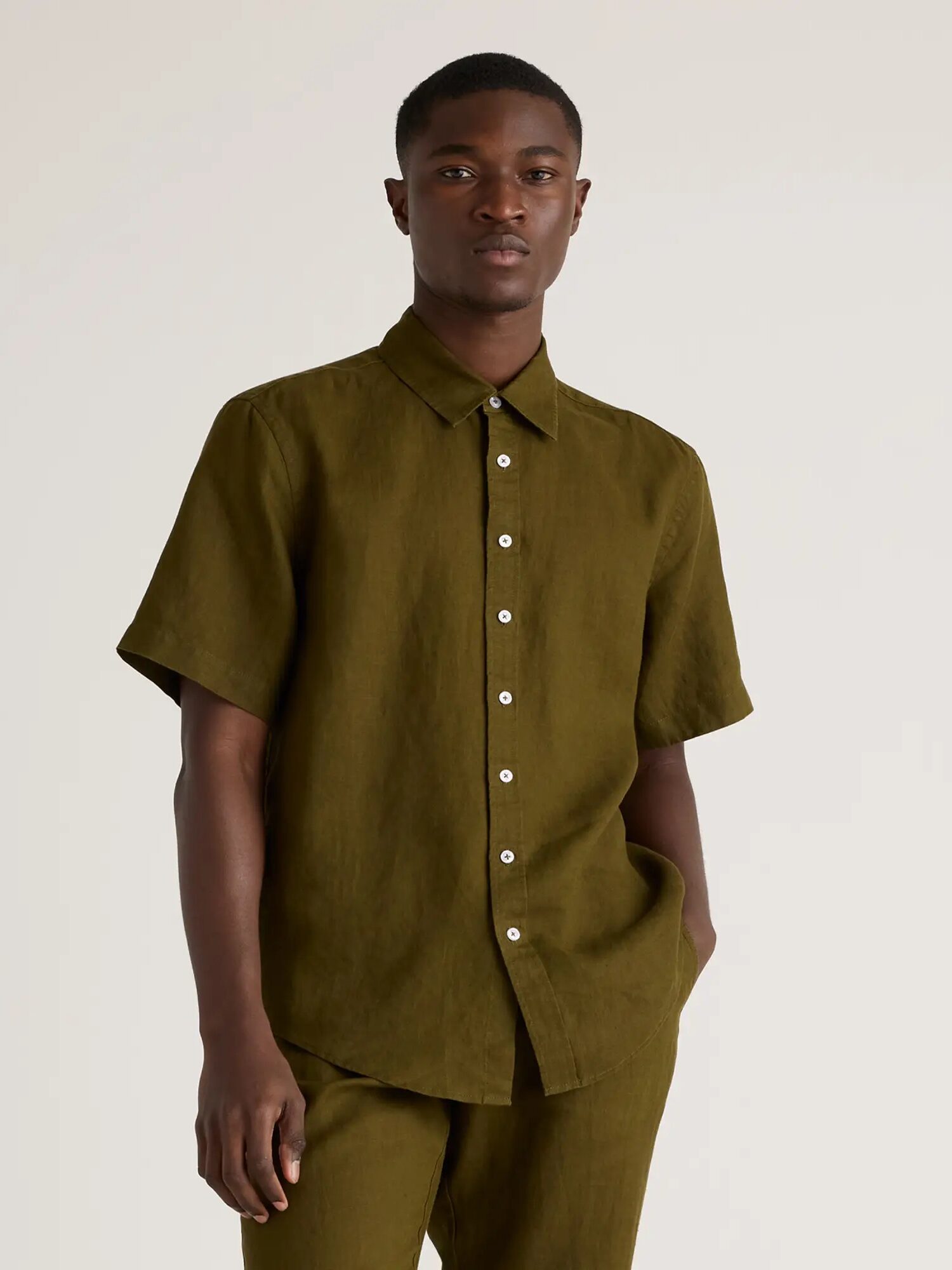 A man wearing a short-sleeve earthy green button-up shirt and matching pants stands in front of a plain background. 