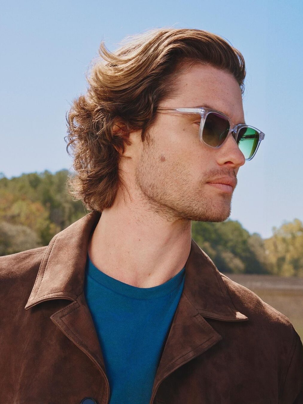 A man with wavy hair wearing mirrored sunglasses, a blue shirt, and a brown jacket stands outdoors with a clear sky and green foliage in the background.