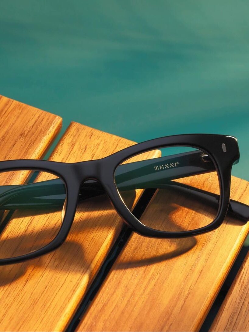 Black-rimmed eyeglasses with a Zenni logo on a wooden surface next to a swimming pool.