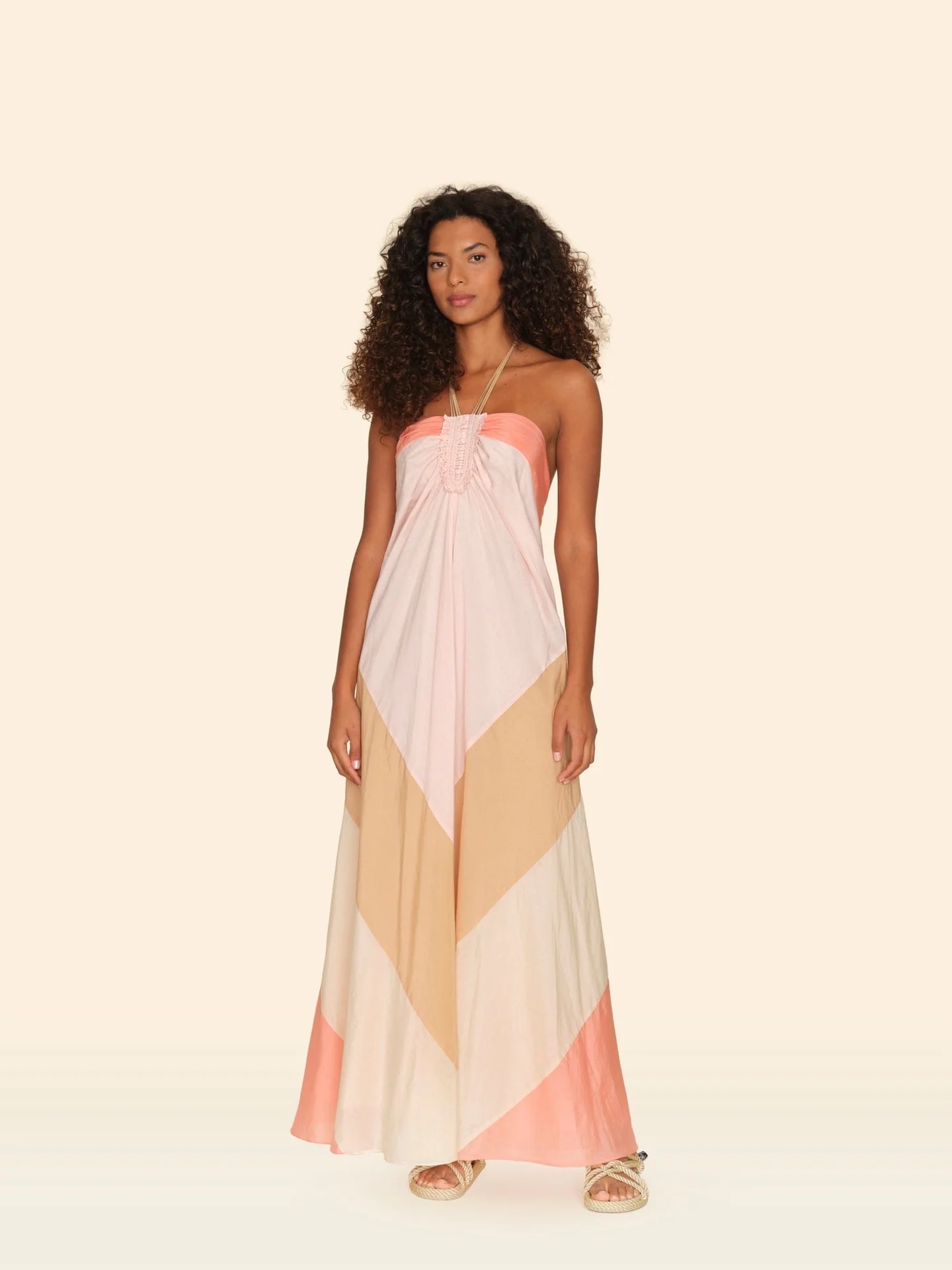 Woman wearing a strapless, geometric-patterned maxi dress in shades of peach and beige, paired with beige sandals, standing against a neutral background.