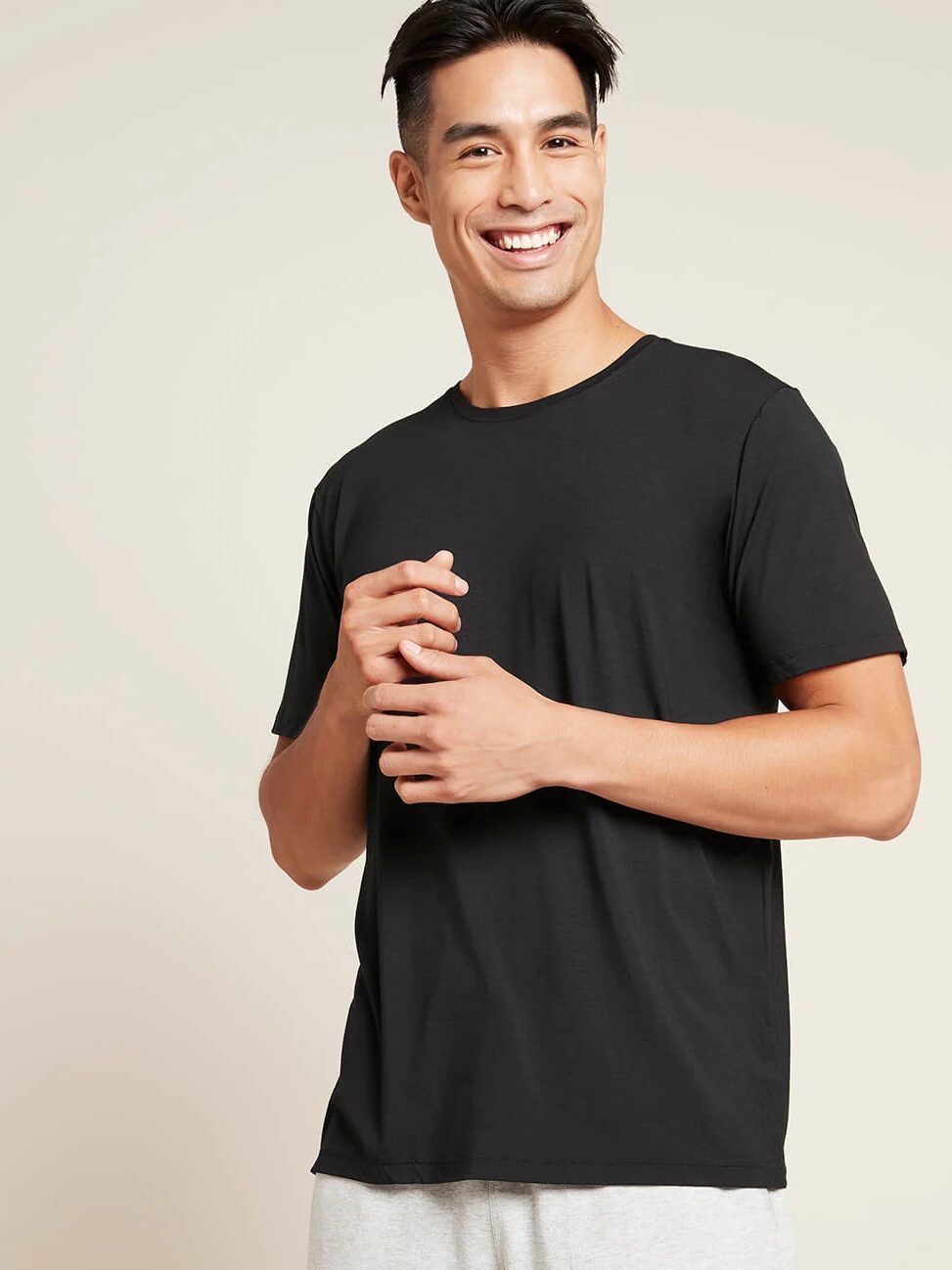 A man in a black t-shirt and grey pants smiles and looks at the camera, standing against a plain beige background.