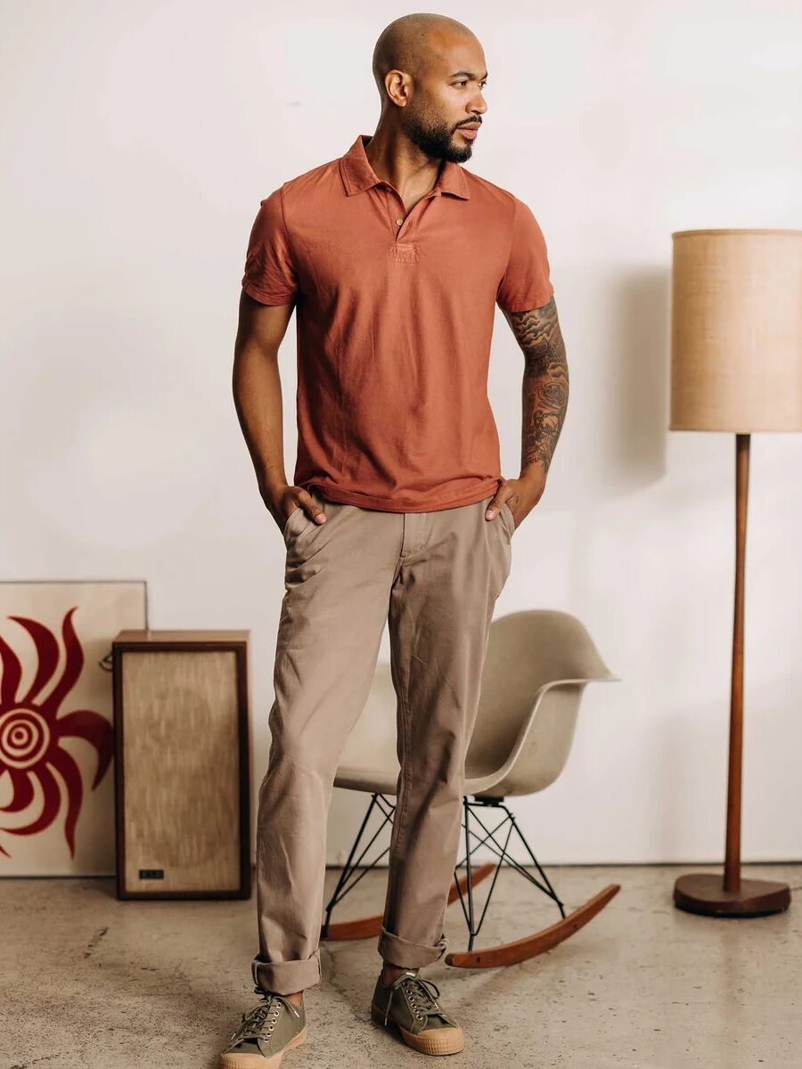A man with a short haircut and a tattoo on his right arm stands in a room with a coral polo shirt and beige pants. There is a decorative artwork, a chair, and a floor lamp in the background.
