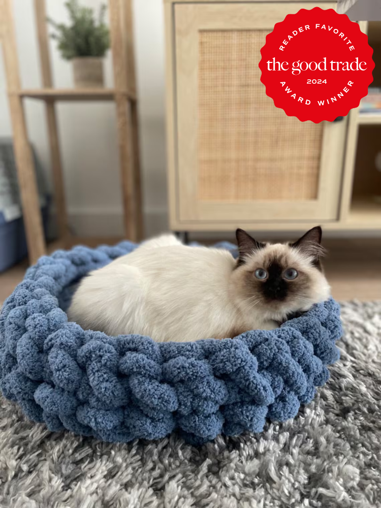 A cat with blue eyes lies in a blue, chunky knit cat bed on a gray carpet. A "Reader Favorite - The Good Trade 2024 Award Winner" badge is displayed in the top right corner.