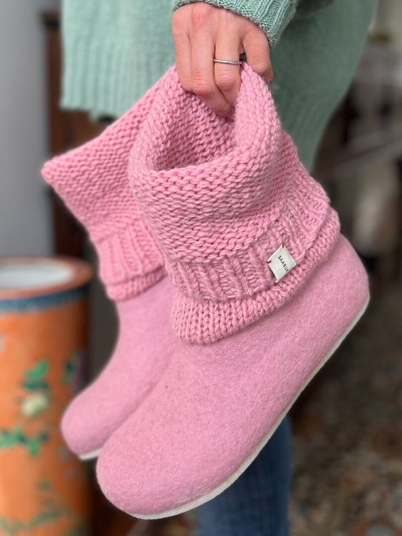 A person holds up a pair of pink knit slippers, which have a rounded, soft sole. The background includes a portion of a green sweater and an orange floral vase.