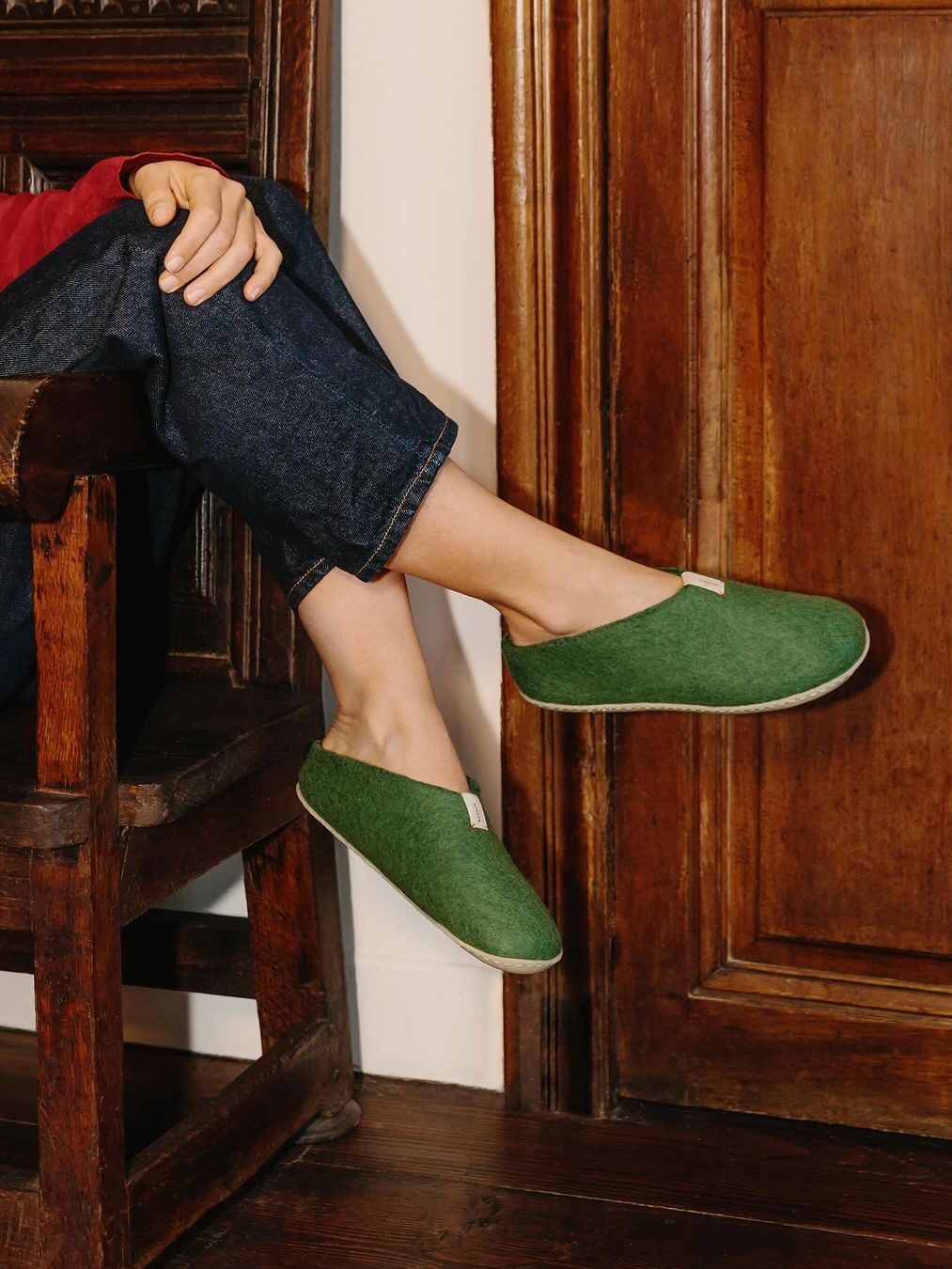 Person sitting in a wooden chair, wearing green slip-on shoes and jeans, with feet resting on a wooden wall.