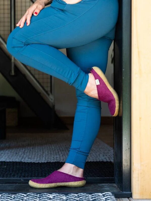 A person in blue pants and purple slippers leans against a wall with one leg crossed over the other, inside a room with a black staircase and a low table in the background.