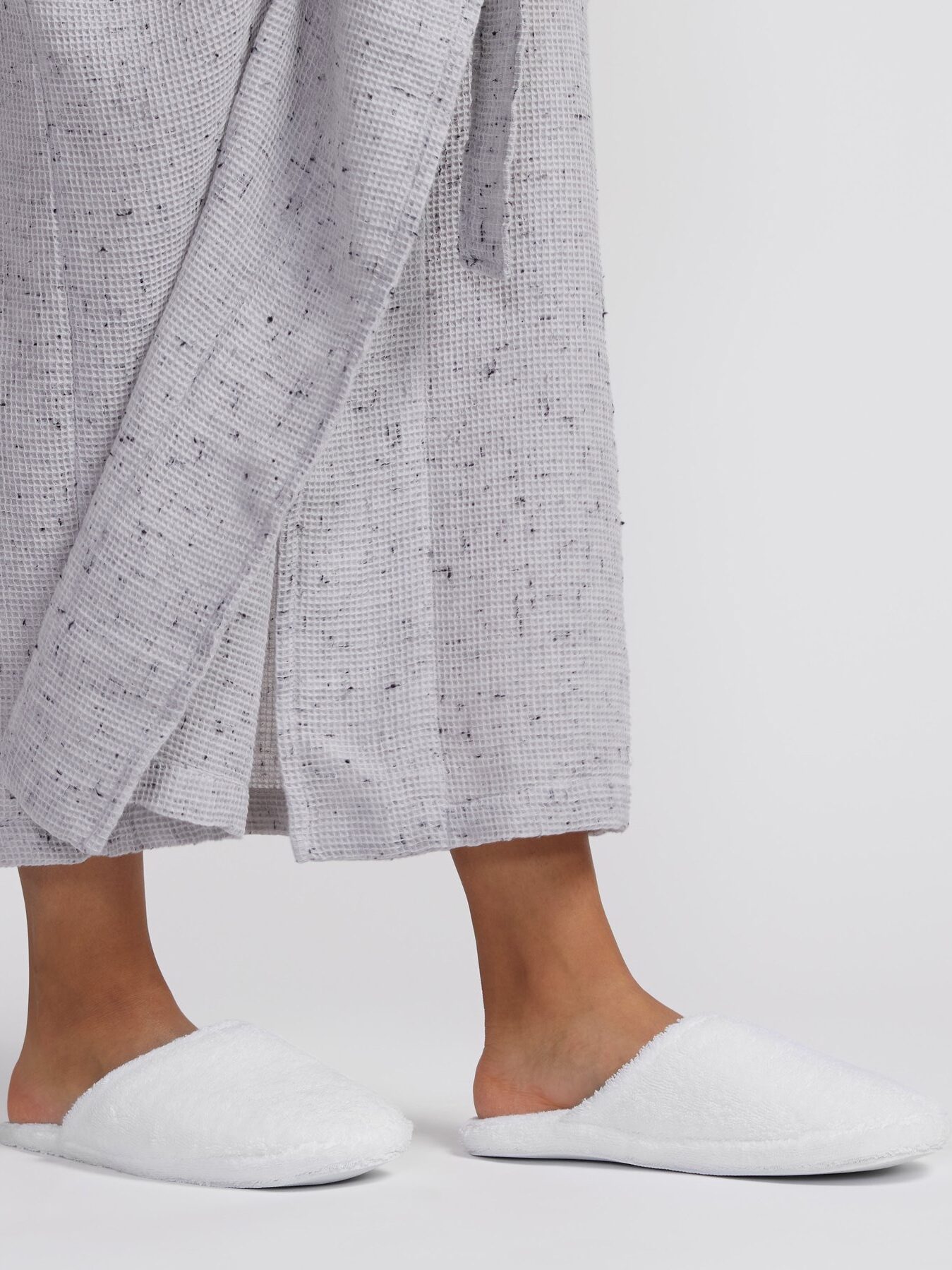 Person wearing a grey textured robe stands on a white background, with their feet in white fuzzy slippers.