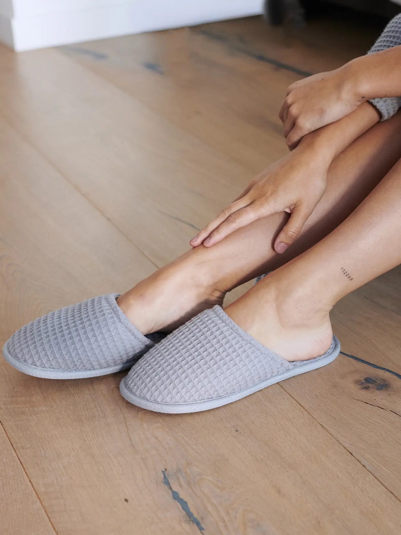 A person sitting on a wooden floor, wearing cozy gray waffle-textured slippers and gently touching their ankle.