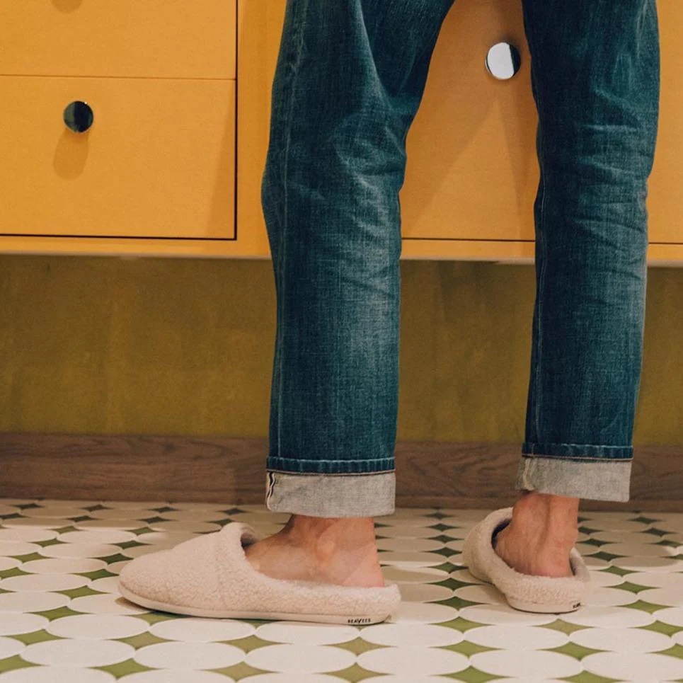 A person in rolled-up jeans and beige slippers stands on a green and white patterned floor in front of a yellow cabinet.