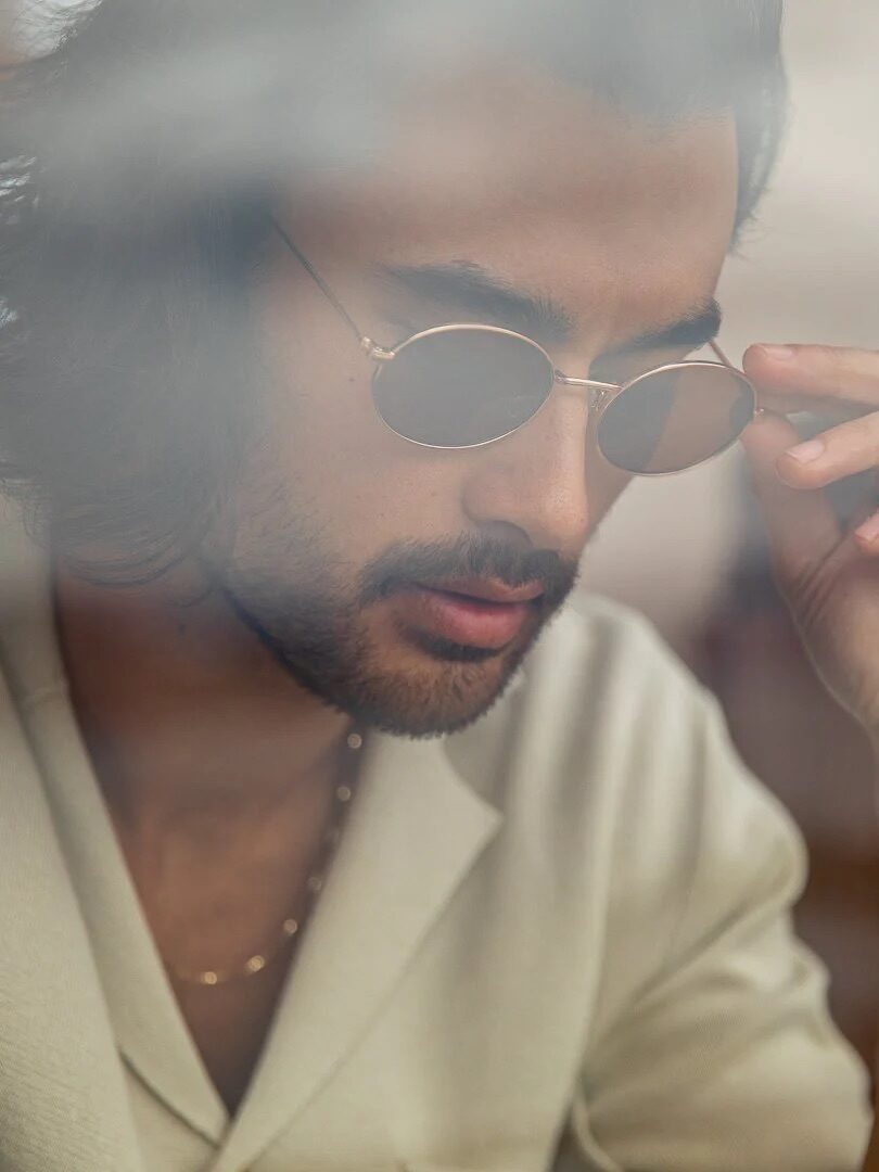 A person with long hair and a beard, wearing sunglasses, a light-colored shirt, and a gold ring, is captured in a candid moment through a foggy lens.