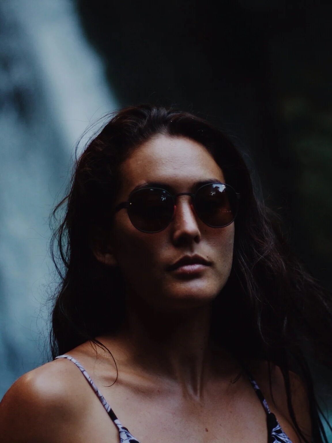 A woman with long dark hair and sunglasses stands in front of a waterfall, wearing a patterned tank top.