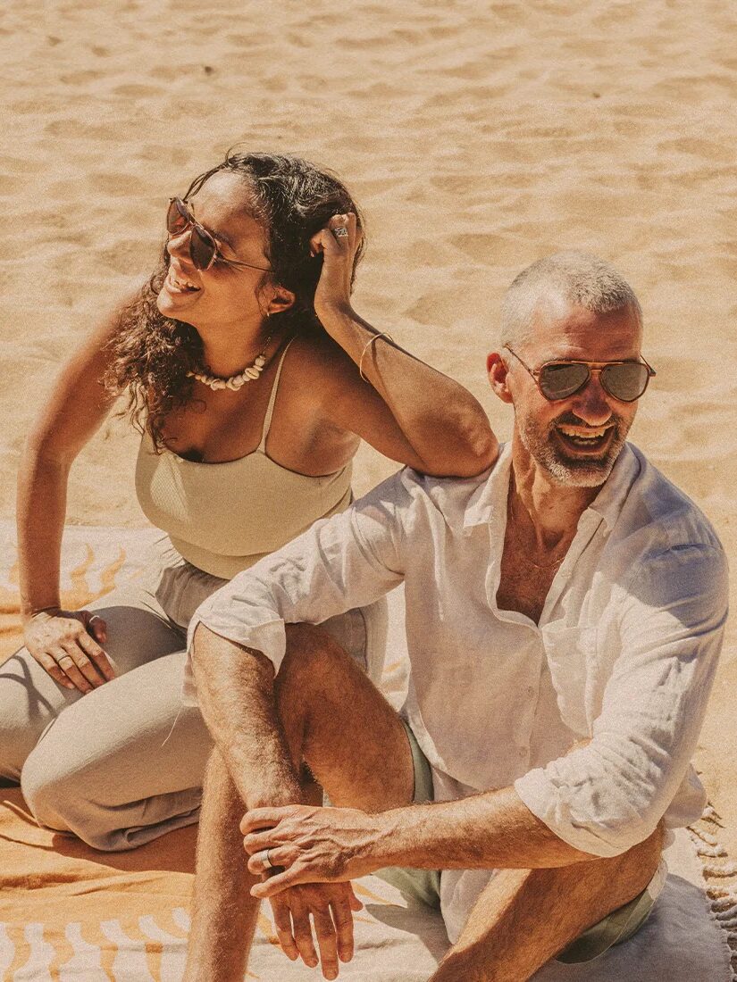 Two people sitting on a beach towel under the sun, both wearing sunglasses and light clothing. There is a wicker bag on the towel.