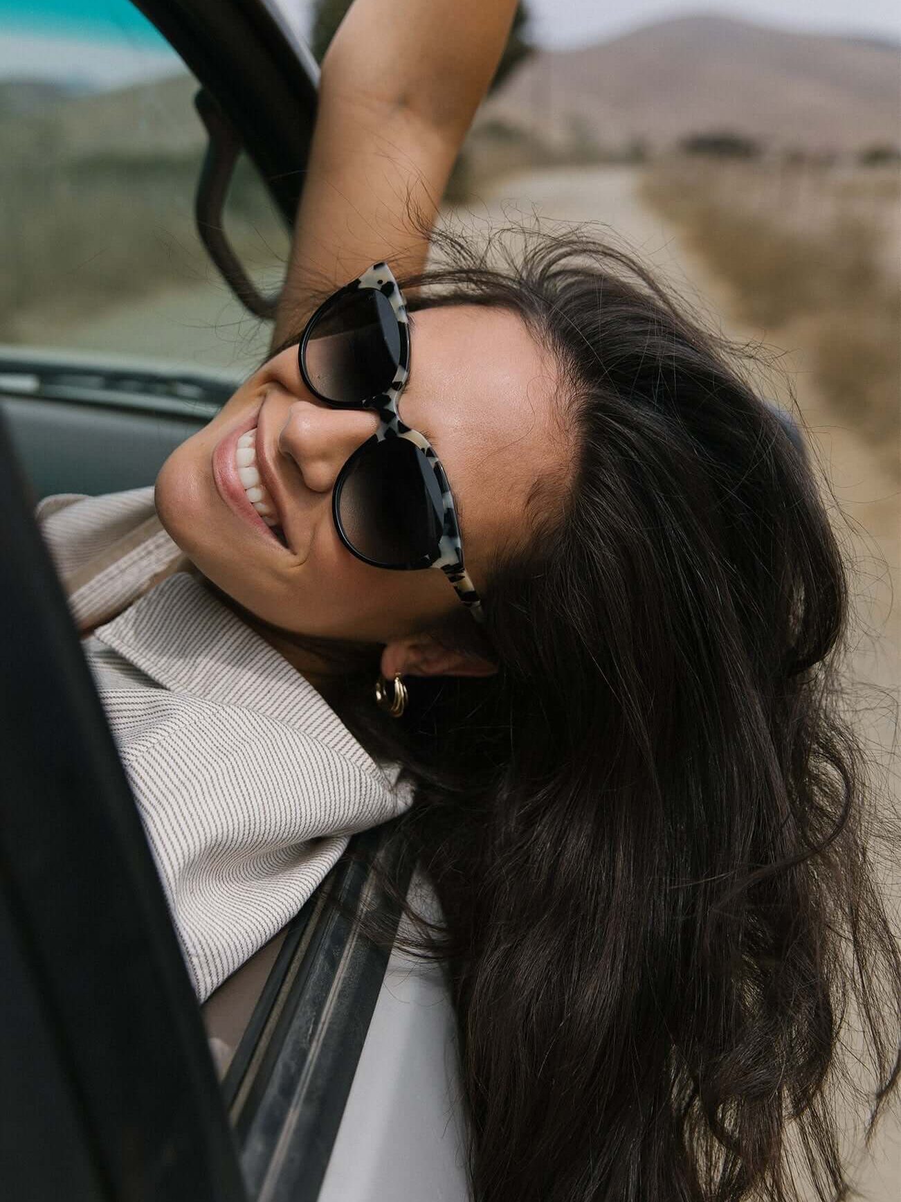 Woman with long hair and sunglasses smiles while leaning out of a car window, with a countryside road in the background.