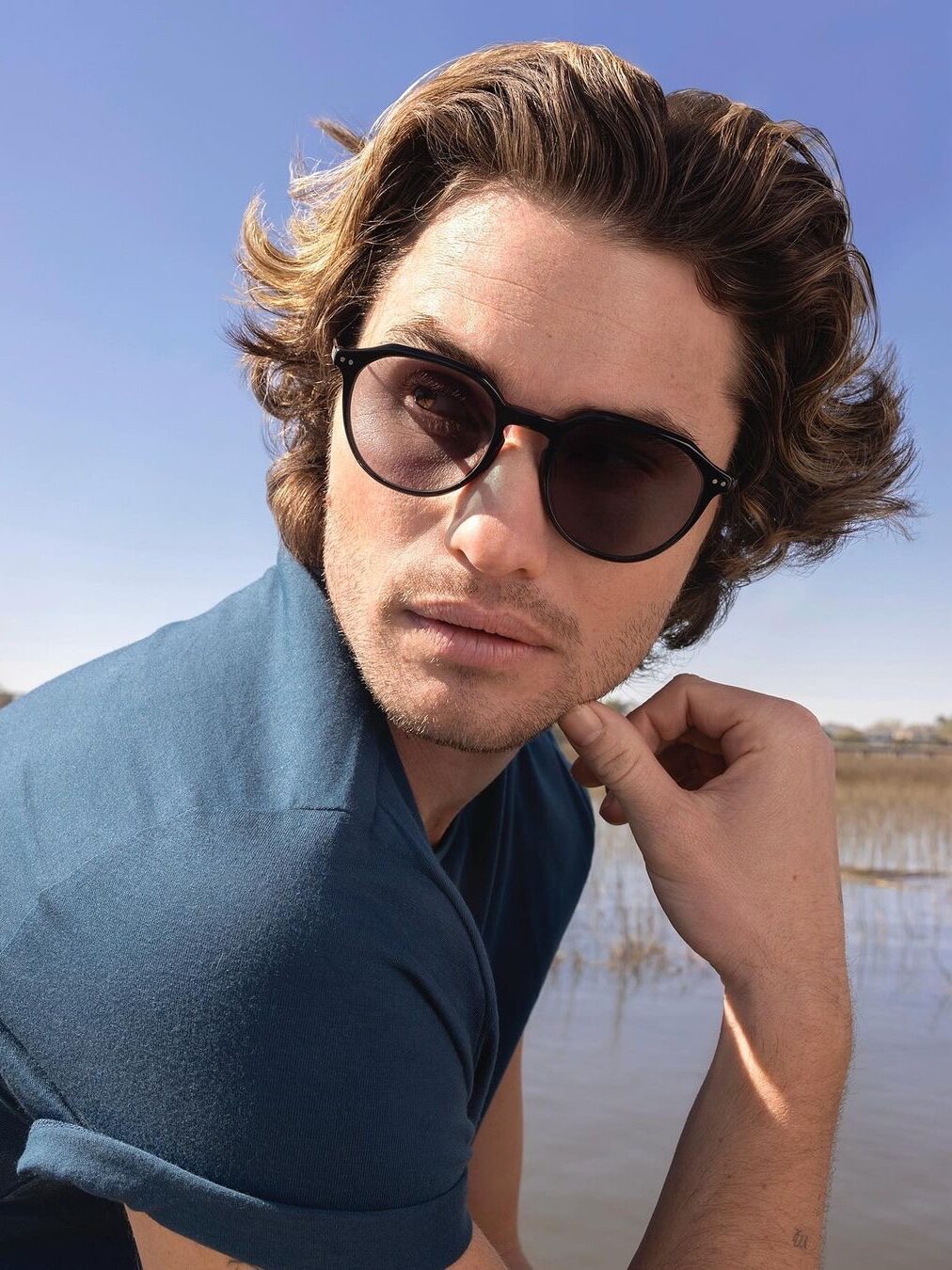 A person with wavy hair wearing sunglasses and a blue shirt looks to the side with a thoughtful expression against a backdrop of water and sky.